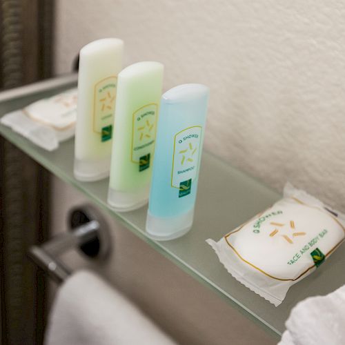 Hotel toiletry items, including three small bottles of shampoo, conditioner, and body wash, and two bars of soap, placed on a glass shelf.