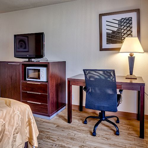 A hotel room with a bed, TV on a cabinet beside a microwave, a desk with a chair and a lamp, and a framed picture on the wall.