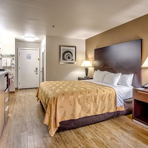 A hotel room with a large bed, TV, desk, lamps, and a picture on the wall above the headboard. Room has wooden flooring.