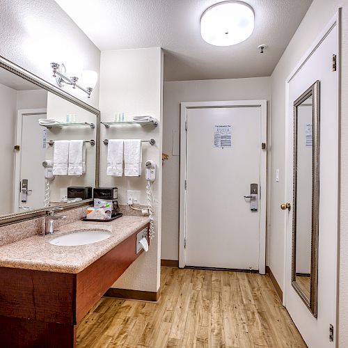 This image shows a hotel room's vanity area with a sink, mirror, toiletries, towels, and a door with a mirror attached to it, under ceiling lighting.