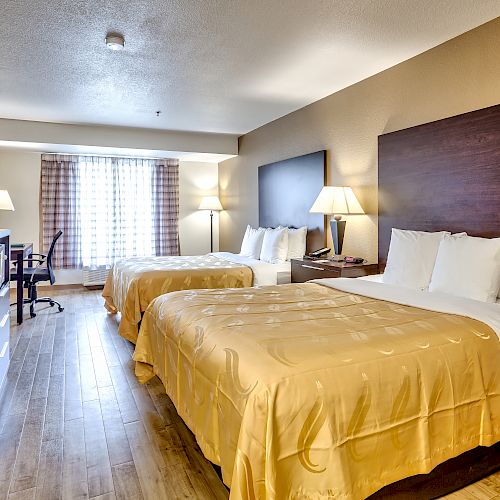 A hotel room with two large beds, wooden floor, desk, flat-screen TV, lamps, and a window with curtains.