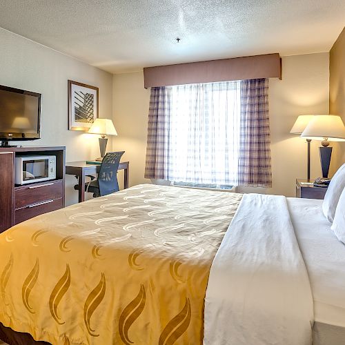 A neatly furnished hotel room features a bed, TV, microwave, desk, chair, and lamps, with a window covered by curtains, offering a cozy ambiance.