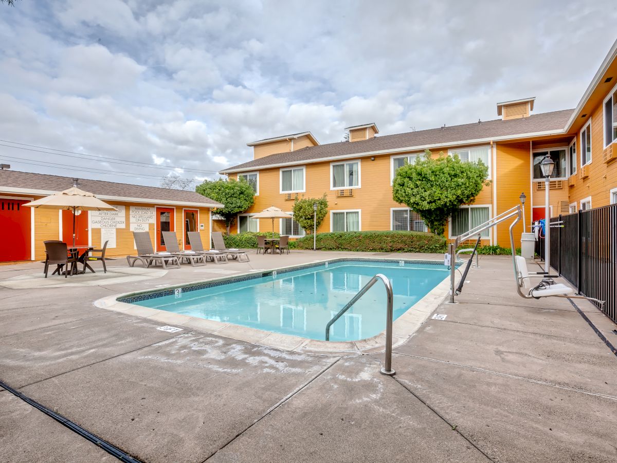 An outdoor pool area at an apartment complex or hotel with lounge chairs, tables with umbrellas, and a two-story building in the background.