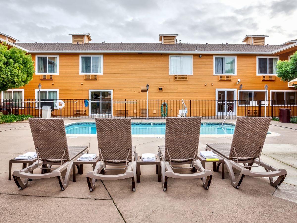 An outdoor pool area featuring four poolside lounge chairs facing a pool, with a building and trees in the background.