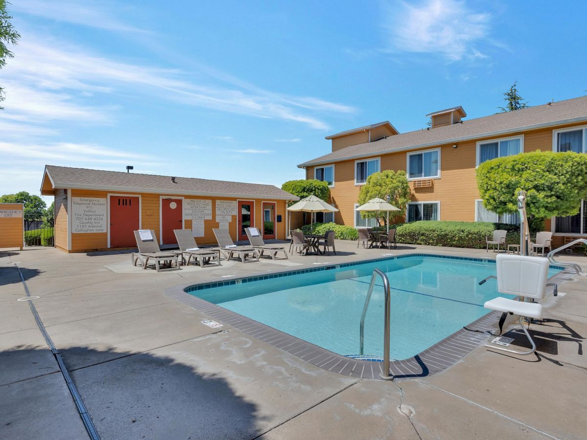 The image shows a hotel pool area with lounge chairs, umbrellas, and a small pool lift, surrounded by a building and trees under a clear sky.