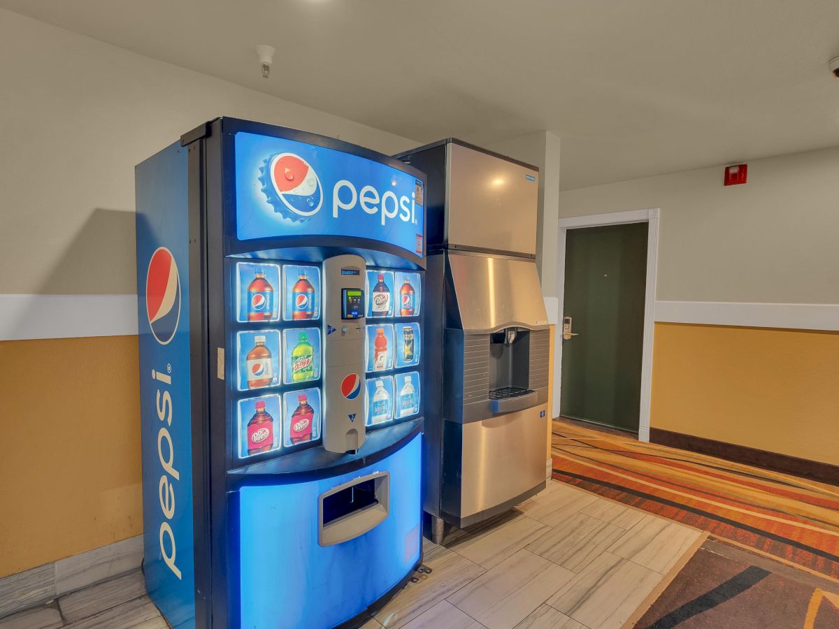 The image shows a blue Pepsi vending machine and an ice dispenser in a hallway with beige walls and a striped floor.