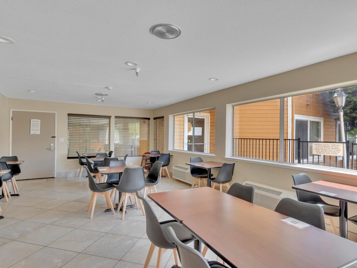 The image shows an empty dining area with several tables and chairs, large windows, and beige walls, creating a bright and clean atmosphere.