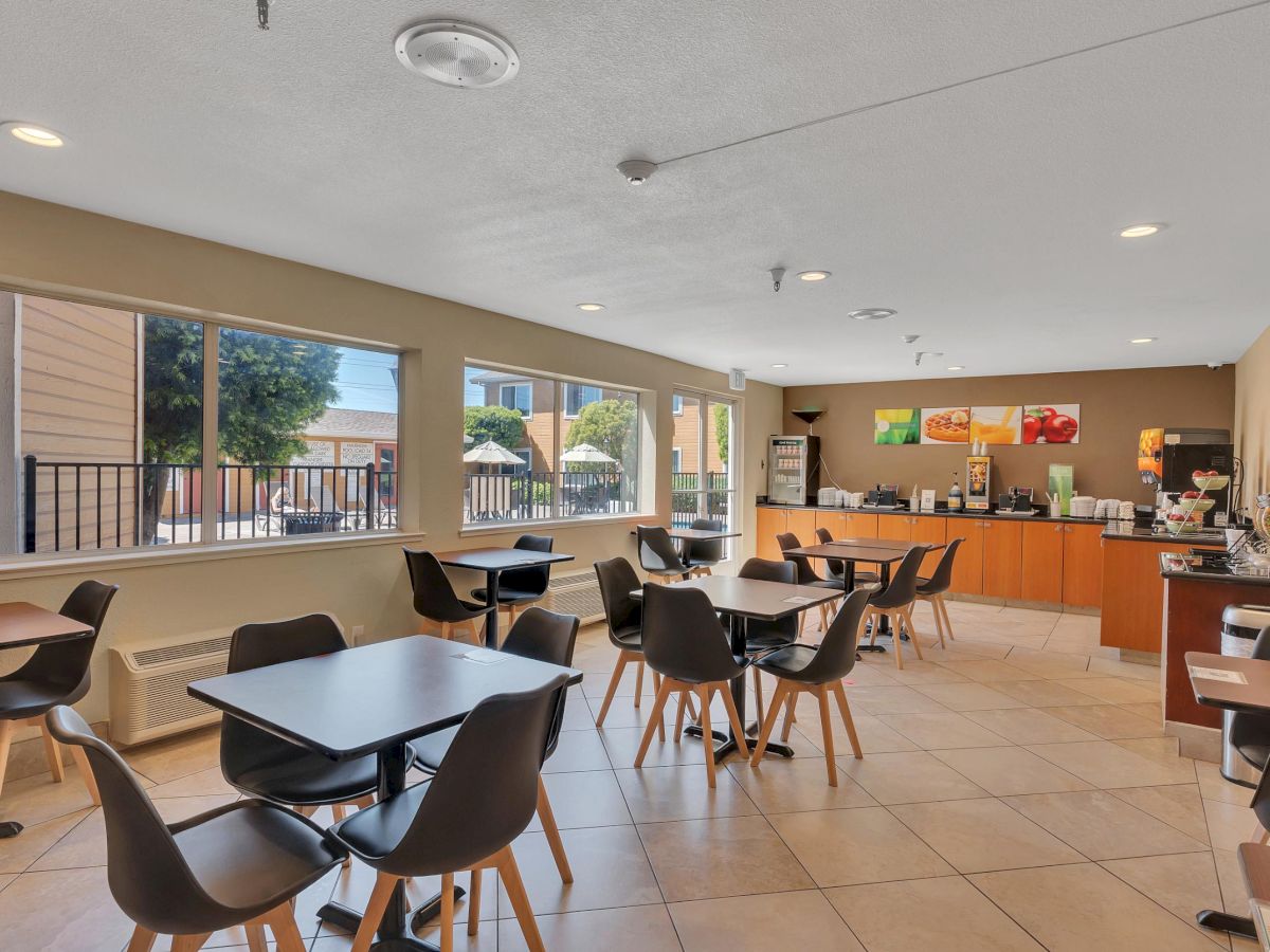 The image shows a dining area with tables, chairs, and a counter setup for food service. Large windows allow natural light to brighten the space.