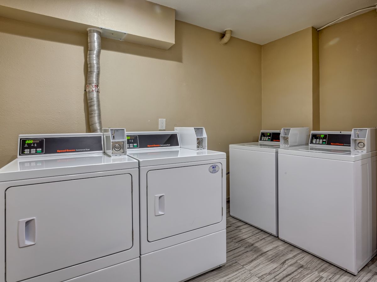 A small laundry room with four white washing machines or dryers, two on each side, against beige walls and a gray floor.