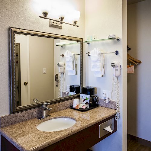 A bathroom vanity with a mirror, sink, toiletries, a coffee maker, and towels on a rack is pictured. There’s a door and a wall hook visible.