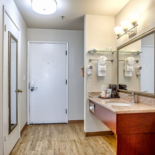 This image shows a hotel or motel bathroom with a large mirror above a vanity, towels, and a well-lit area with a door and a closet.