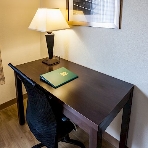 A small desk with a black chair, a table lamp, a framed picture, and a closed green binder is situated in the corner of a room.