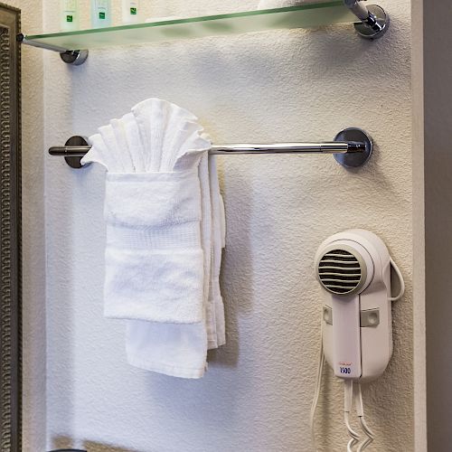 The image shows a bathroom setup with a glass shelf holding toiletries, a towel rack with folded towels, a hairdryer, and an outlet near a coffee maker.