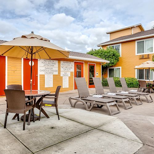 The image shows an outdoor area with lounge chairs, tables, and umbrellas near a building with several windows and doors, under a partly cloudy sky.