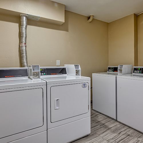 The image shows a laundry room with four coin-operated washing machines and dryers placed against beige walls on a wooden floor.