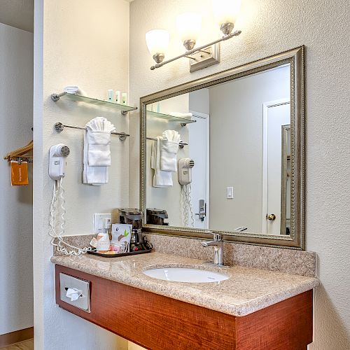 A bathroom vanity with a mirror, sink, towel rack, wall-mounted hairdryer, and toiletries on the counter, under bright lighting, is shown.