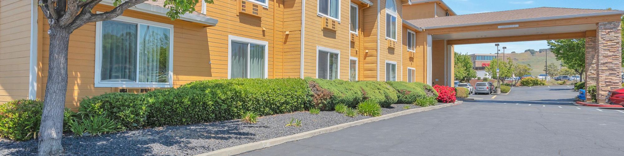 The image shows a two-story building with a yellow facade, surrounded by greenery, and a driveway with a covered entrance.