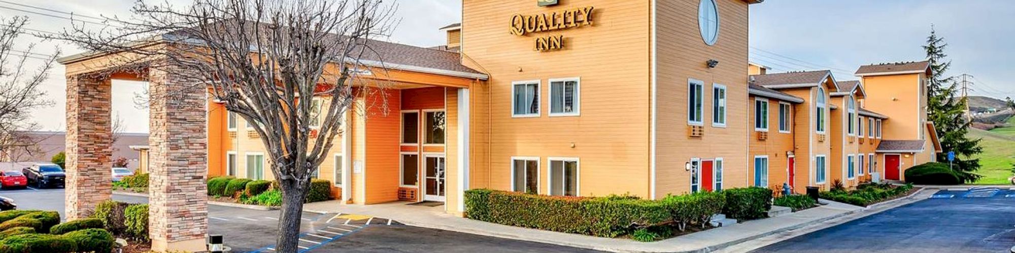 A two-story hotel with a parking lot in front, labeled "Quality Inn," surrounded by minimal landscaping and trees under a partly cloudy sky.