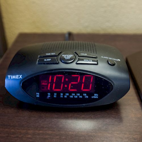 A Timex digital alarm clock displaying the time as 10:20 is on a wooden nightstand next to a brown lamp.