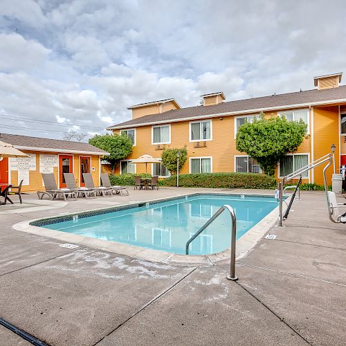 The image shows an outdoor swimming pool area surrounded by a beige building with multiple windows and doors, a patio umbrella, and lounge chairs.