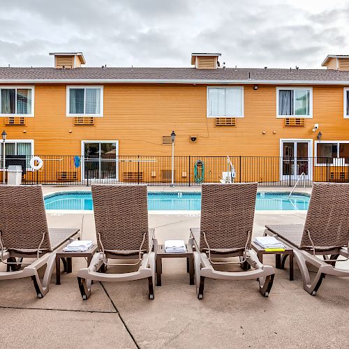 Four lounge chairs facing a pool with a two-story yellow building in the background and trees on either side.