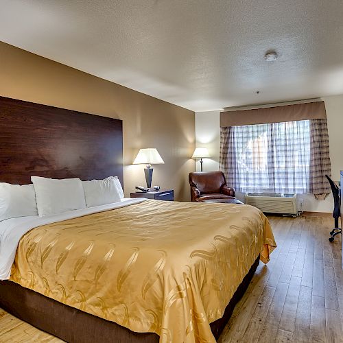 A cozy hotel room with a large bed, two side lamps, a leather chair, a wooden dresser with a TV, and a window with checkered curtains.