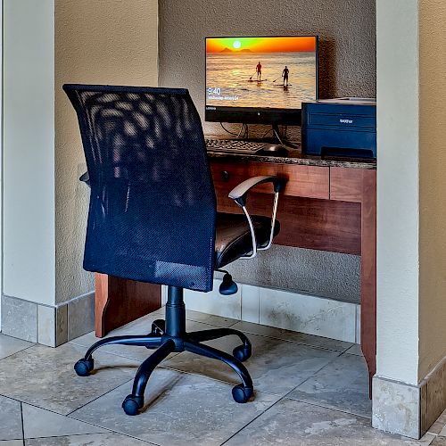 A small office setup with a computer on a desk, and a black chair in front of it. The monitor shows a beach scene with a sunset.