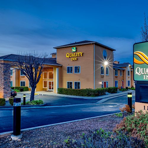 The image shows a Quality Inn hotel with its sign lit up at dusk, featuring the main entrance and surrounding landscaping.