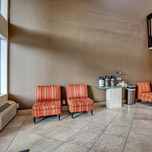 The image shows a seating area with striped chairs, a table with coffee and tea dispensers, and tiled flooring. The area has a high ceiling and large window.