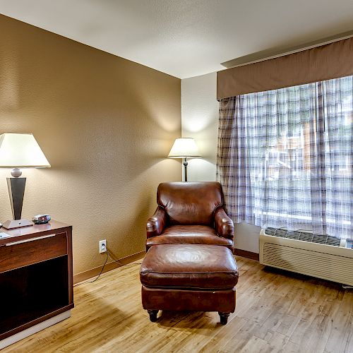 The image shows a cozy room with a leather armchair and ottoman, side table with lamp, and a window with plaid curtains. There's also an AC unit.