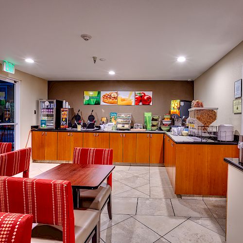 The image shows a small breakfast area with tables and chairs, a counter with breakfast items, a coffee machine, and various food dispensers.