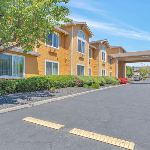 A yellow two-story building with a covered entrance, surrounded by landscaping and a paved driveway, under a clear blue sky.