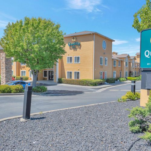 The image shows a hotel entrance with a sign reading "Quality Inn" surrounded by trees and a neatly landscaped area.