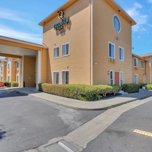 The image shows a two-story hotel building with a beige exterior and green sign, surrounded by a parking lot and landscaped bushes.