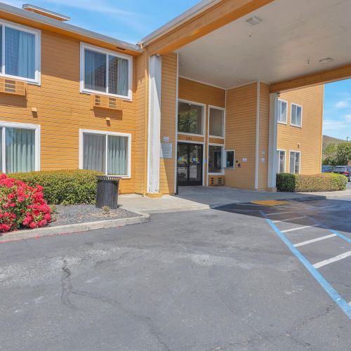 The image shows the entrance of a two-story building with tan siding, windows, red bushes, and a covered driveway.