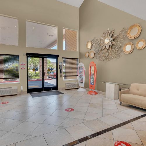 The image shows a spacious lobby with modern decor, a beige couch, wall art, large windows, and safety markers on the floor.