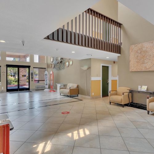 The image shows a spacious lobby with chairs, red partitions, tiled floors, and large windows allowing natural light to enter.
