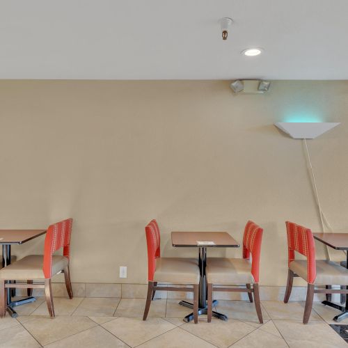 The image shows a simple interior with two small tables and red chairs against a beige wall, with a wall-mounted light above.