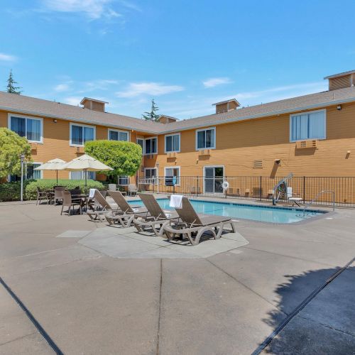 The image shows a courtyard with a pool, lounge chairs, and umbrellas, surrounded by a two-story building with windows and trees.