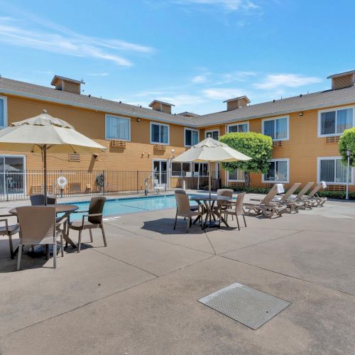 A courtyard features a swimming pool, surrounded by lounge chairs, tables with umbrellas, and a two-story building in the background.