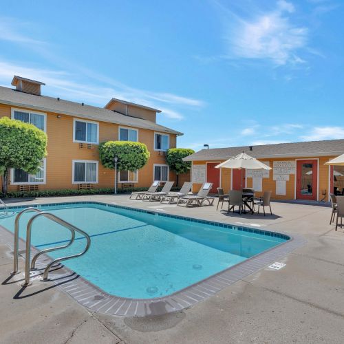 The image shows an outdoor swimming pool area with loungers, umbrellas, and a two-story building in the background on a sunny day.