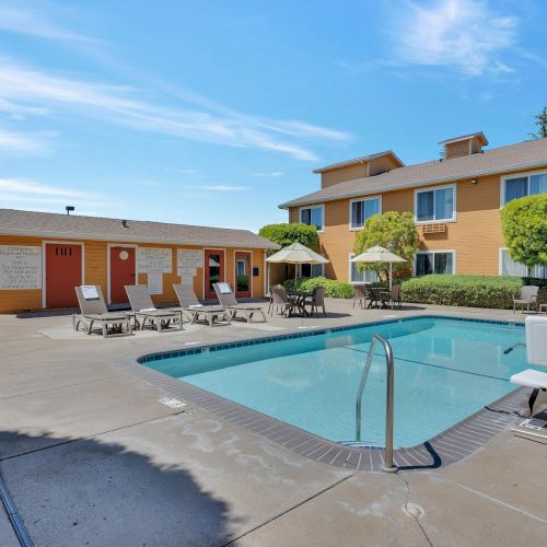 The image shows an outdoor swimming pool area with lounge chairs and umbrellas, next to a building with trees and clear skies above.