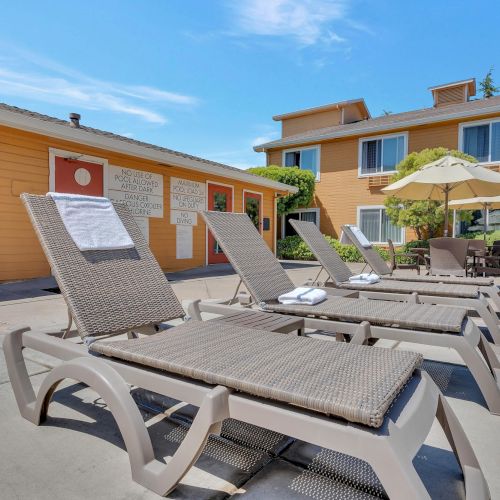Poolside area with several lounge chairs, a couple of buildings, and a patio umbrella in a sunny setting.