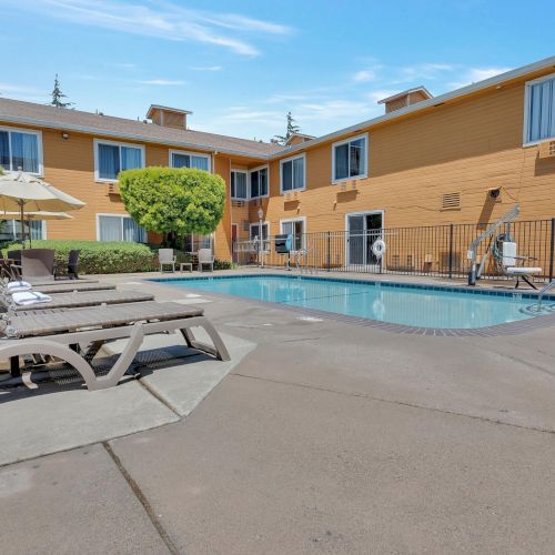 A small outdoor pool area with lounge chairs and umbrellas, surrounded by a two-story building under a clear sky.