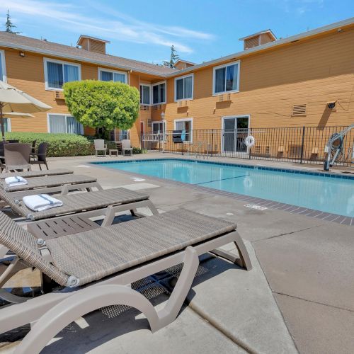The image shows a hotel courtyard with a swimming pool, lounge chairs, and a beige building in the background under a clear blue sky.