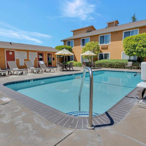 The image shows an outdoor swimming pool with lounge chairs and umbrellas surrounded by buildings under a clear blue sky.