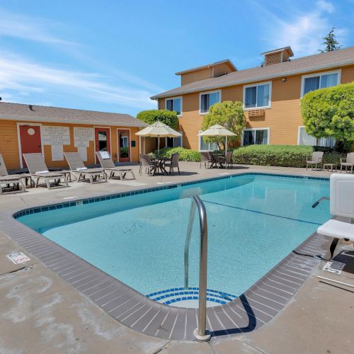 The image shows an outdoor pool area with lounge chairs, umbrellas, and a building in the background under a clear blue sky.