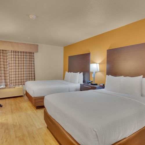 The image shows a hotel room with two double beds, a nightstand with a lamp, a desk and chair, and curtains over the window.