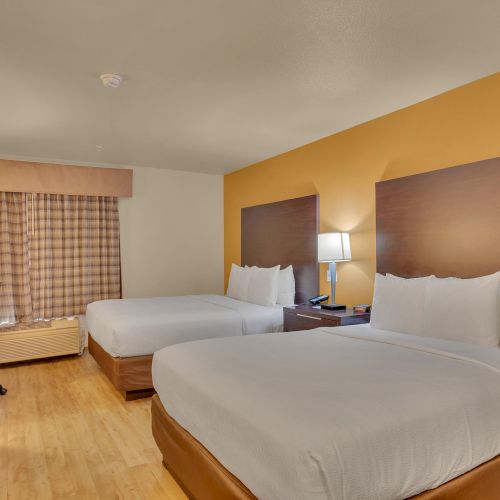 The image shows a hotel room with two double beds, a nightstand with a lamp, a desk with a chair, and a TV.