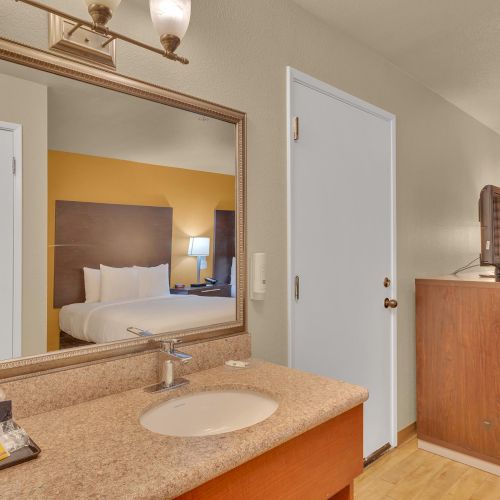 The image shows a hotel room with a bed, a bathroom sink, a mirror, a desk with a chair, and some soft lighting.